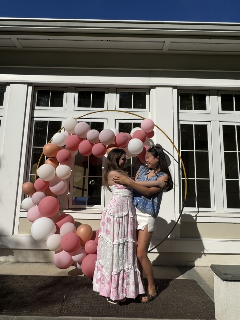 Two friends laughing under a pink balloon arch